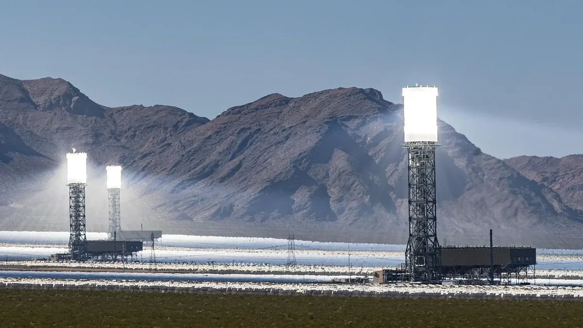 A solar thermal power plant with two large towers emitting bright light, situated in a desert landscape with mountains in the background.