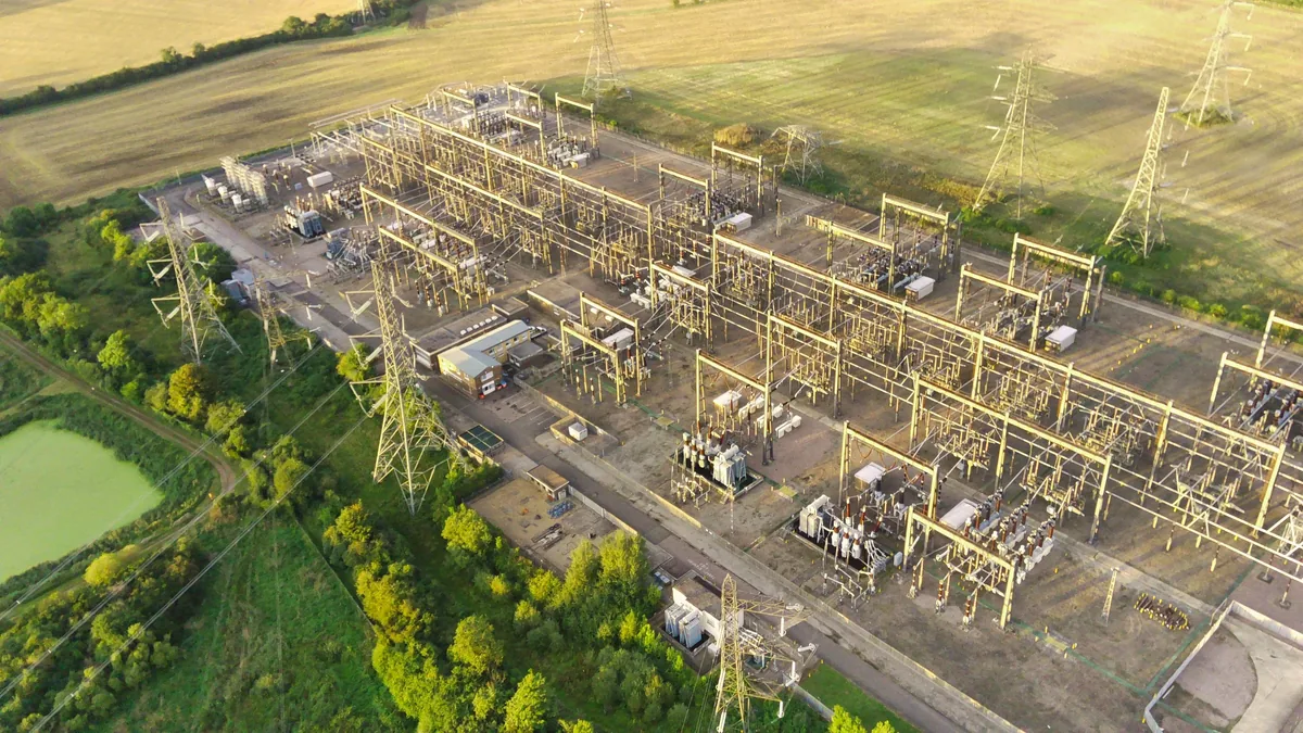 A large electrical substation with numerous power lines and equipment, situated in a rural area with fields and trees.