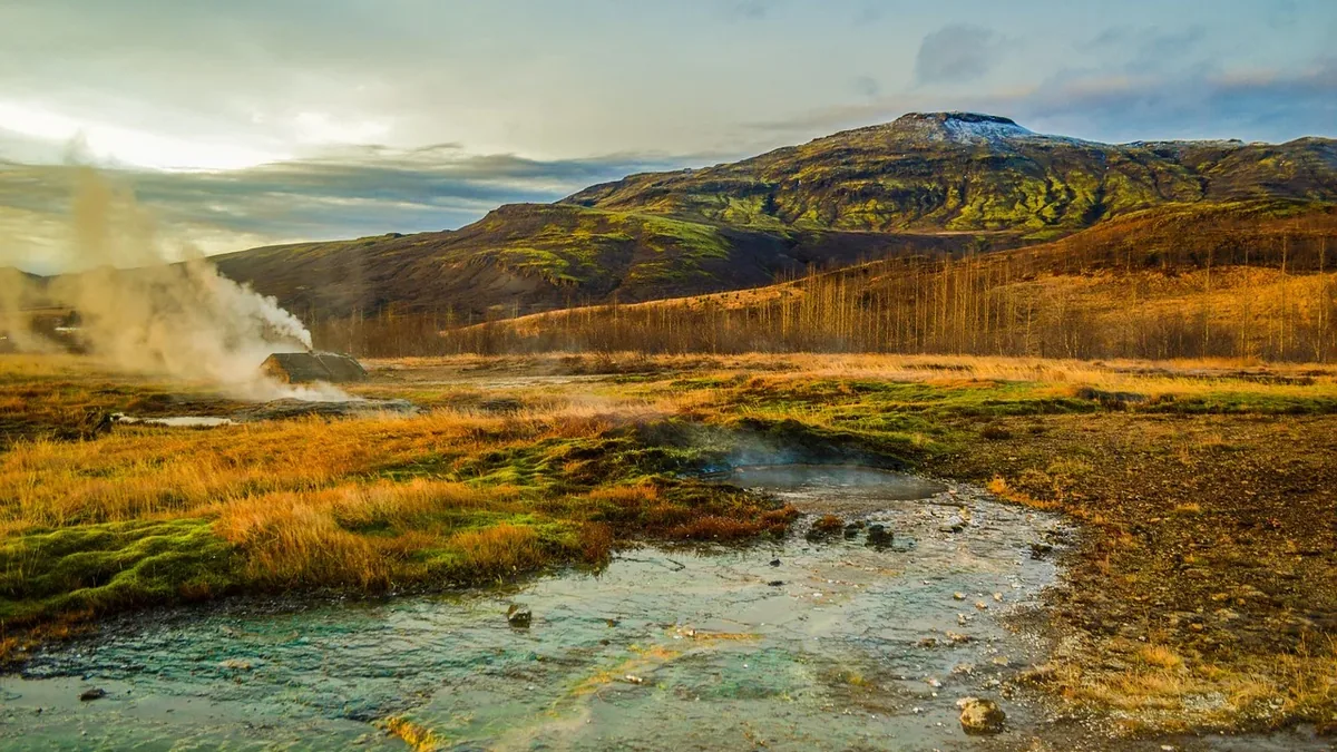 A geothermal area with a stream of water, steam rising from the ground, and a mountainous landscape in the background.