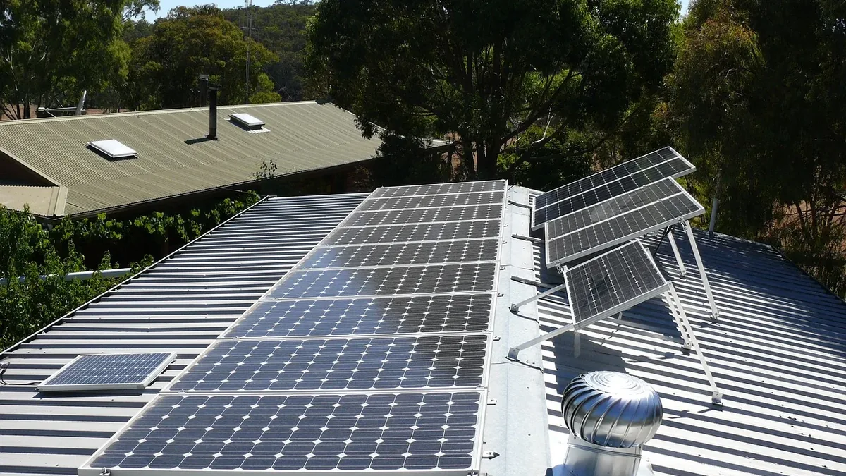 A rooftop with solar panels and a ventilation system, set against a backdrop of trees and a house.