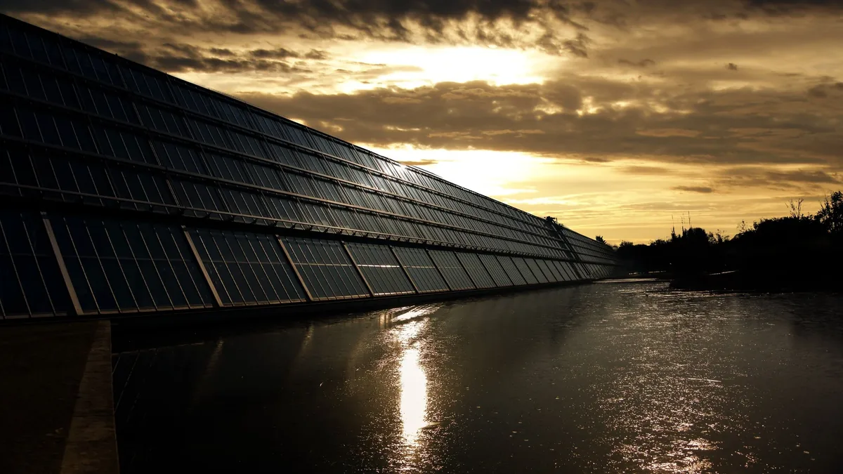 A long row of solar panels installed on a bridge over a body of water, with the sun setting in the background.